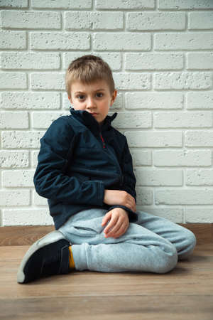 A pensive boy of 6-7 years old sits on the floor with a mysterious look. Portrait of a boy, front viewの写真素材
