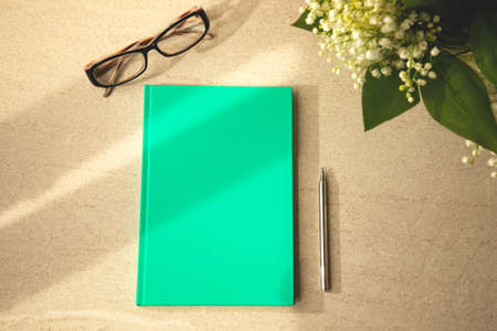 Green notebook on a brown table with a pen, glasses and a bouquet of lilies of the valley. Office...