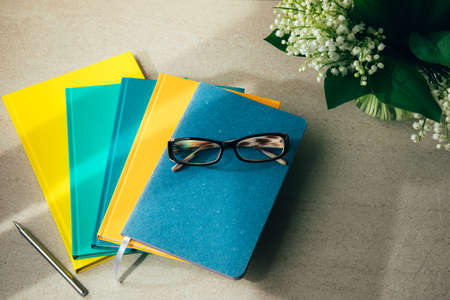 Multi-colored office notebooks on a light beige background and glasses on the top with a bouquet of lilies of the valley. Set of notebooks, front viewの写真素材