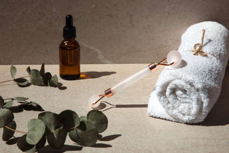 Rose quartz facial massage roller and white towel on a stone table with eucalyptus branches and a bottle of serum. Front viewの写真素材