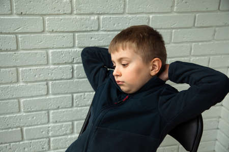 A pensive boy of 6-7 years old sits on a chair on the balcony and looks out the window. Portrait of a boyの写真素材