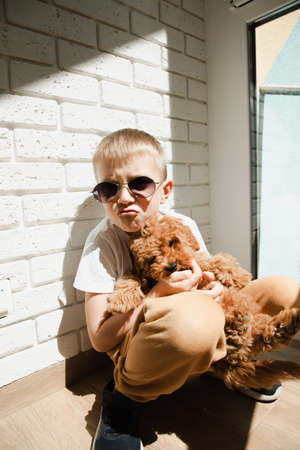 A boy in sunglasses sits near a brick wall and smiles with a puppy. Portrait of a boy 6-7 years old