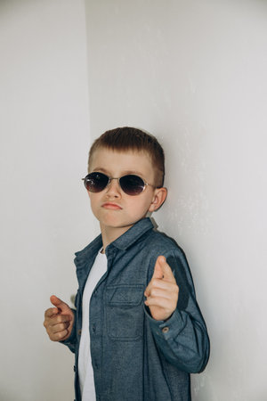 A cool boy stands dressed in a shirt peeking out from under sunglasses
