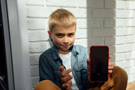 Boy 6-7 years old in a blue shirt sitting on the floor near the window pointing at the smartphone screenの写真素材