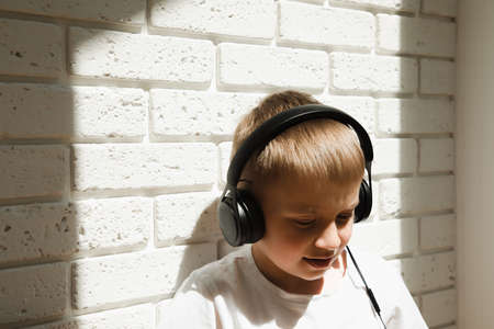 A boy of 6-7 years old in a white T-shirt listens to music in black headphones and looks downの写真素材