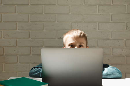 Schoolboy is sitting at the office desk and peeking out from behind a gray laptop near the windowの写真素材