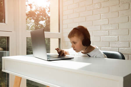 Boy 6-7 years old at a white table near the window in large headphones uses a laptopの写真素材