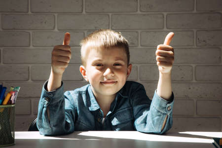 A schoolboy sits at a white desk in a blue shirt and shows a thumbs up gesture. Front viewの写真素材