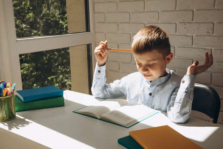 A schoolboy holding a yellow pencil near his head sitting at a white desk. Back to school concept