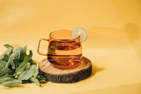 Mint tea in a glass orange mug on a wooden stand on a yellow background with a shadow. Front viewの写真素材