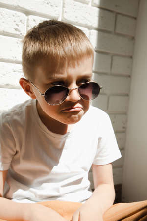 A boy in sunglasses sits leaning against a brick wall and grimaces. Portrait of a boy 6-7 years old