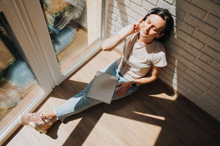 A woman is sitting on the floor near the window wearing headphones and a laptop on her feet and listening to music with eyes closed. View from aboveの写真素材