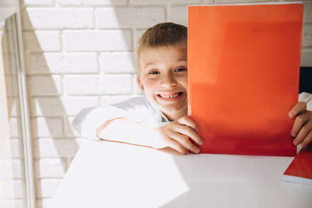 A boy in a white shirt at a white table shows a red textbook with a place for writing and looks from behind the book. Back to school conceptの写真素材