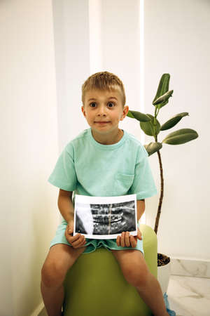 A 6-7-year-old boy is sitting on a green chair in a dental clinic with a picture of his teeth in his hands. Portrait of a boy with an x-rayの写真素材