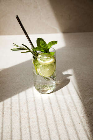 Drink in a glass with mint, rosemary, lemon and lime on a stone table with shadow. Summer drink lemonade with an iron tubeの写真素材