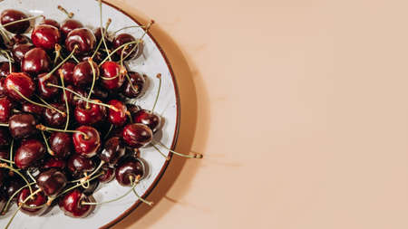 Banner of a large cherry close up in a white deep plate on a light orange background in sunlight. Top viewの写真素材