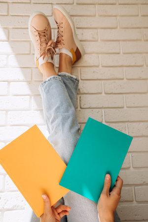 Woman holding an two close books or notebooks with her legs up in blue jeans and pink sneakers against a brick wall backgroundの写真素材