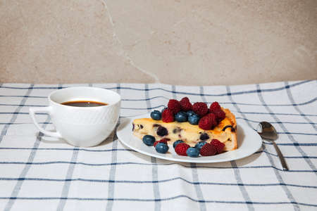 A piece of blueberry cheesecake decorated with raspberries and blueberries on the kitchen table with a cup of coffee. Front viewの写真素材