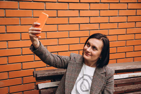 A girl in a brown jacket sits on a bench and takes a selfie. Portrait of a dark-haired girl against...