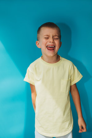 A 6-7-year-old boy in a yellow T-shirt on a blue background shouts loudly. Portrait of a child, front viewの写真素材