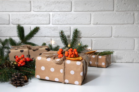 Group of gifts in eco packaging, wrapped in brown paper with white circles and decorated with a dry orange, mountain ash and a fir branch. New Years gifts in lights on white table, front viewの写真素材