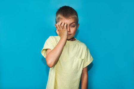 A 6-7 year old boy in a yellow T-shirt shows a gesture of being bored covering his face with one hand on a blue background. Close-up portrait, front viewの写真素材