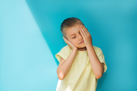 A boy in a yellow T-shirt on a blue background smiling covers his face with his hands. Portrait of a sleepy, tired child, front viewの写真素材