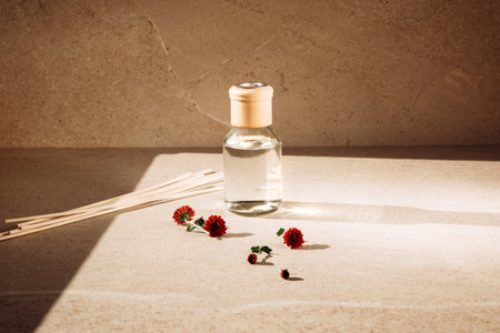 Aroma diffuser in a clear glass bottle with a wooden cap on a light stone background with wooden sticks behind and red chrysanthemum flowers. Front viewの写真素材