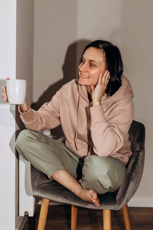 A girl in a pink hoodie sits on a gray chair with a cup of tea in her hands and looks out the window. Front viewの写真素材