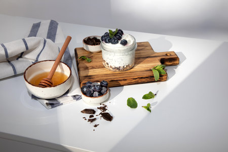 Yogurt in a glass jar with granola, chocolate, blueberries and honey on a wooden board on a white background with mint leaves. Top viewの写真素材