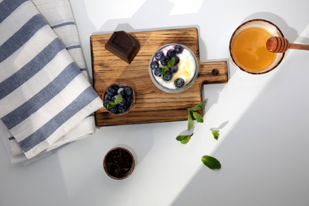 Yogurt in a glass jar with chocolate, blueberries and honey on a wooden board on a white background with mint leaves. Flat layの写真素材