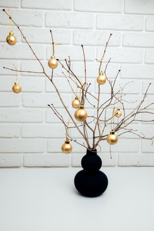 Christmas decoration, dry tree branches in a black vase decorated with golden balls against a white brick wall. Front viewの写真素材