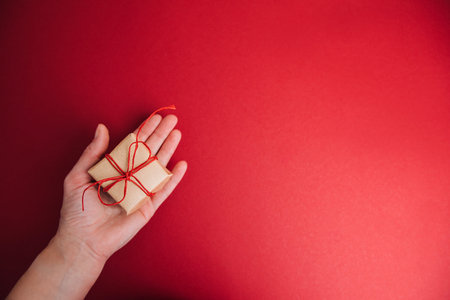 A gift wrapped in brown paper with a red ribbon on a red background is held by a female hand. Christmas gift, flat layの写真素材
