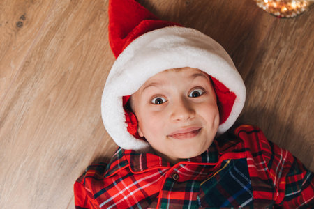 A joyful boy in red-green pajamas and a Santa hat lies on the floor. Christmas at home. Front viewの写真素材