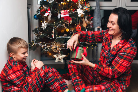 Mom and son in red pajamas sit near the Christmas tree and exchange gifts. Front viewの写真素材