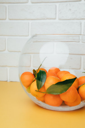 Orange mandarins clementine with green leaves in round clear vase on a white-yellow background. Front viewの写真素材