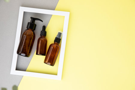 Amber spray and dispenser bottles with facial cosmetics in the white frame on the grey-yellow background. Empty space for design. Flat layの写真素材
