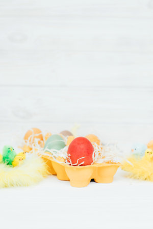 Painted multi-colored eggs in a yellow cardboard tray on a white wooden background. Easter...