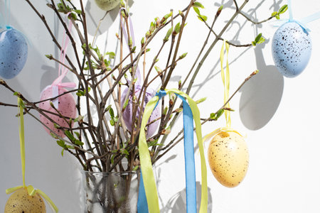 Tree branches with young green leaves in a transparent vase are decorated with colorful Easter eggs with shadows on a white background. Front viewの写真素材