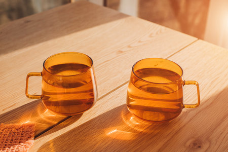 Two orange transparent glass mugs with hot tea with yellow reflection on a wooden table in the kitchen in sunlight. Front viewの写真素材