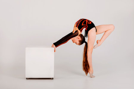 A girl gymnast in a black leotard does tricks on a white cube on a white background. Front viewの写真素材