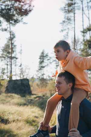 Dad and son on a walk in the forest in sunny weather. Family leisure. The father carries his son on his shoulders. Front viewの写真素材