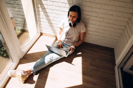 A woman is sitting on the floor near the window wearing headphones around the neck and a laptop on her feet. Top viewの写真素材