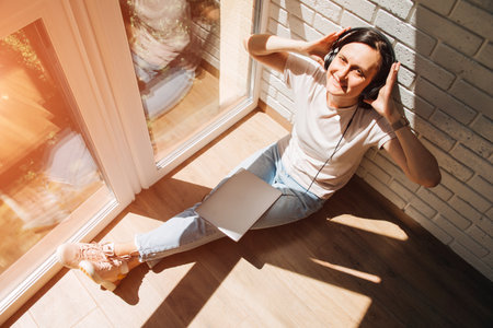 A woman is sitting on the floor near the window wearing headphones around the neck and a laptop on her feet. Top viewの写真素材