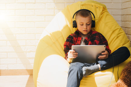 A school-age boy in black headphones sits on a yellow bean bag with a tablet in his hands. Children and gadgets. Front viewの写真素材