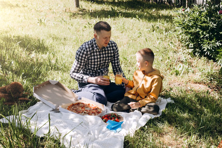 Family picnic. Dad and son in a green garden in sunny weather on a picnic eating pizza and drinking juice. Front viewの写真素材