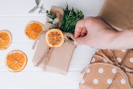 The gift is wrapped by female hands in brown paper and tied with twine with dry orange and eucalyptus on a white wooden background. Top viewの写真素材