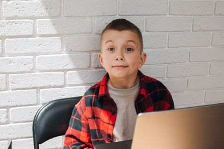A smiling school-age boy sits at a table with a laptop. Children and gadgets. Front view