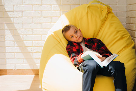 A school-age boy sits on a yellow bean bag with a book in his hands. Children reading. Front viewの写真素材