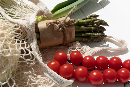 Set of fresh vegetables, asparagus, tomatoes, onion in beige mesh bag on white wooden background....